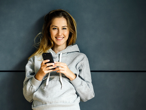 Photo of a beautiful young blond woman leaning up against a wall, smiling while using her cellphone.