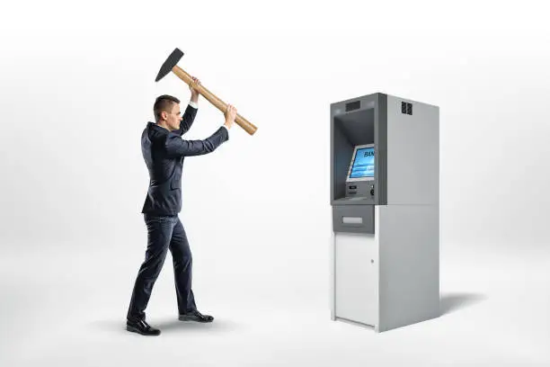 Photo of A businessman holds a heavy hammer high over a working bank ATM machine.