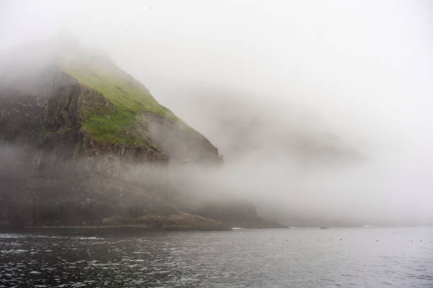 Misty landscape of Mykines island shoot from water. Fantastic green cliffs above the ocean and thick morning fog. Misty landscape of Mykines island mykines faroe islands photos stock pictures, royalty-free photos & images