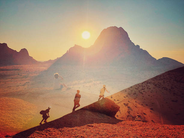 Overlay toned image of a boy sitting on a rock high above the ground overlooking Spitzkoppe as the sun sets and the silhouette of three boys with backpacks walking up a steep hill one with a headlamp flashlight at dusk in Namibia Africa
