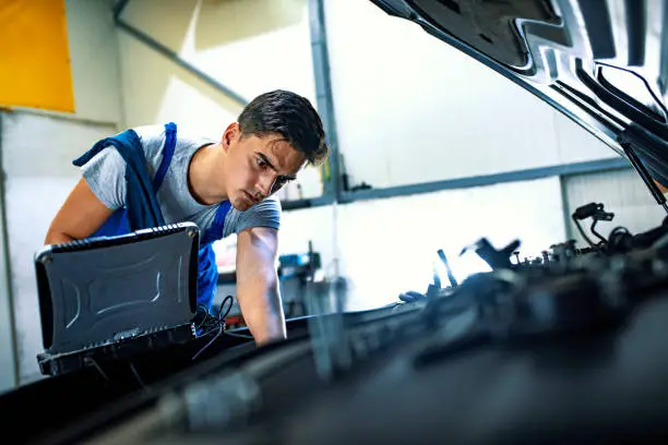 Serious Handsome mechanic in uniform is using a laptop while repairing car in auto service. Mechanic Using Laptop While Examining Car Engine. Skilled mechanic using a laptop computer to check a car engine.