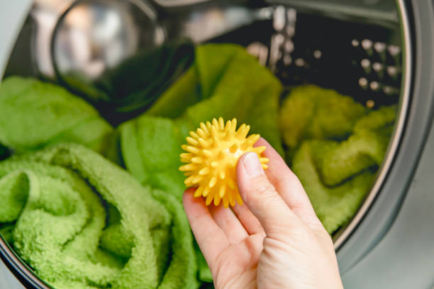 Using pvc dryer balls is natural alternative to both dryer sheets and liquid fabric softener, balls help prevent laundry from clumping in the dryer. Woman hand put in a yellow spiky dryer ball. el uso de bolas de secadora de pvc es una alternativa natural tanto a las sábanas de secadora como al suavizante de tela líquida, las bolas ayudan a evitar que la ropa se agrupe en la secadora. mujer a mano puesto en una bola de secador espinosa amarill