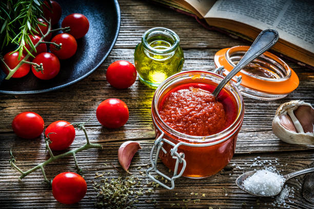 High angle view of homemade tomato sauce in a glass jar shot on rustic kitchen table. Some ingredients for preparing tomato sauce like fresh ripe tomatoes, rosemary, olive oil, peppercorns and salt are all around the tomato sauce jar. DSRL studio photo taken with Canon EOS 5D Mk II and Canon EF 100mm f/2.8L Macro IS USM.