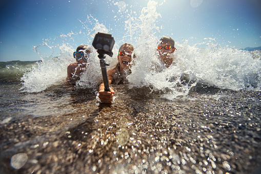 Brothers and sister are having fun in sea. Kids are lying on the front and being splashed by waves. Kids are holding a waterproof action camera on a selfie stick and filming themselves.
Nikon D850