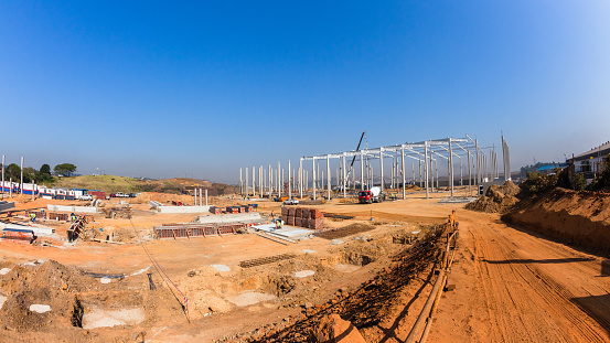 Industrial construction on site of new warehouse factory buildings been erected with concrete columns rigging attaching steel roof beams on structure on a blue sky day landscape.