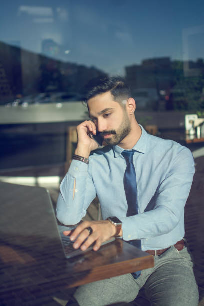 istock Beautiful young middle eastern business man talking on phone while using laptop in cafe. Through window view. 1165336839