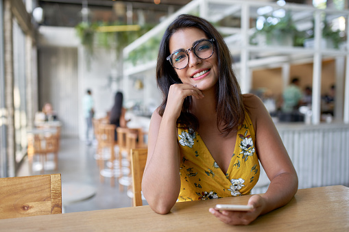 Unposed lifestyle potrait of young charming spanish brunette business woman in her 20s typing on her smartphone, in a bright hipster cafe with many plants and vegetation wearing a yellow floral dress