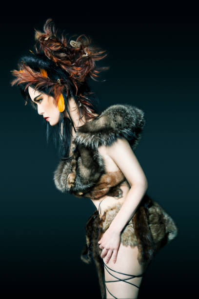 Beautiful Asian woman wearing her black hair up with brown feathers. Wrapped up in fur and laces, bending over. Creative make-up and a fierce look. Looking away, serious expression.