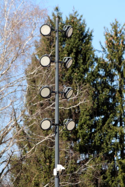 istock Multiple modern LED reflectors and small security camera mounted on tall metal pole at local park surrounded with dense trees 1165357125