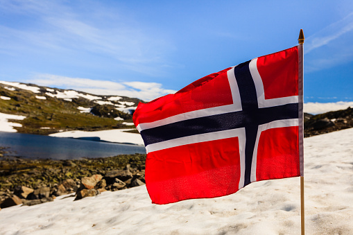 Norwegian flag waving against snowy mountains landscape, summertime. National tourist route Aurlandsfjellet.