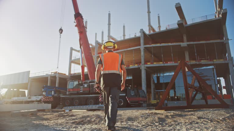 Following Shot: Worker Contractor Wearing Hard Hat and Safety Vests Walks on Industrial Building Construction Site. In the Background Crane, Skyscraper Concrete Formwork Frames