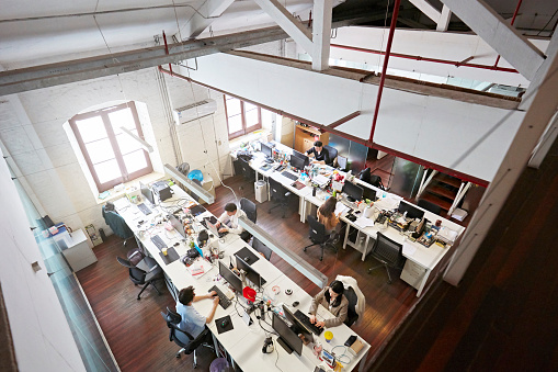 High angle view of small design business office space with staff sitting at long workstations in front of desk top computers.
