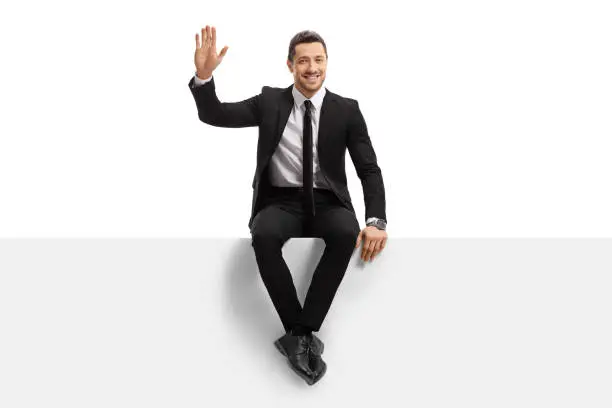Full length portrait of a young man in a suit sitting on a panel and waving at the camera isolated on white background