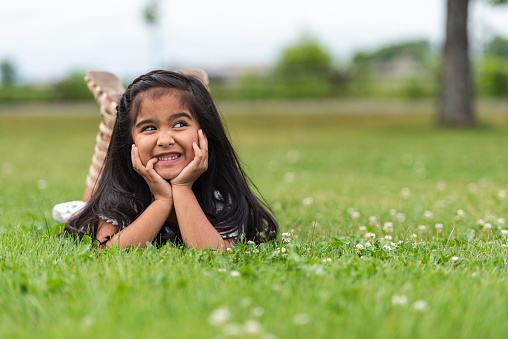 A cute little kindergarten girl of Indian descent lays in the grass with her head in her hands. She is smiling and her legs are up in air. She is bathed in sunlight.