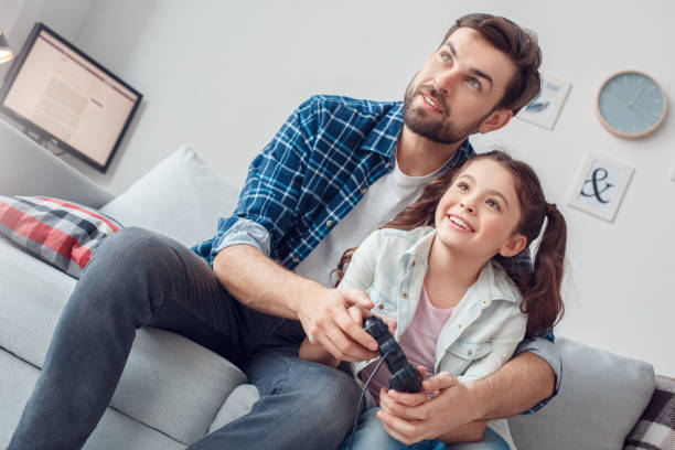istock Father and little daughter at home sitting holding controller together cheerful 1165422583