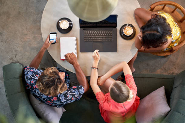 Candid overhead shot of three multi-ethnic millennial coworkers collaborating over coffee with laptop computer at bright cafe serving fair-trade coffee stock photo