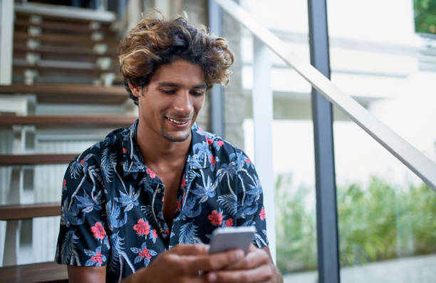 istock Attractive hispanic hipster millennial influencer smiling and texting on cellphone sitting on modern stairs of bright room with tropical plants 1165423269