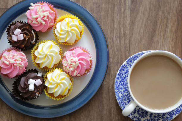 Image of small homemade cupcakes / mini muffins with rainbow multicolour icing piped cakes as flower swirl with orange teapot and glass mug cup of tea, pink, yellow lemon, chocolate icing cakes displayed on round white plate for afternoon tea party buffet