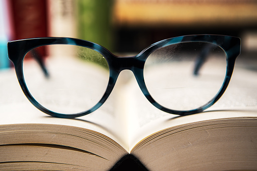 Slightly smudged Eyeglasses with blue frame on open book with blurred books in background. Reading learning concept