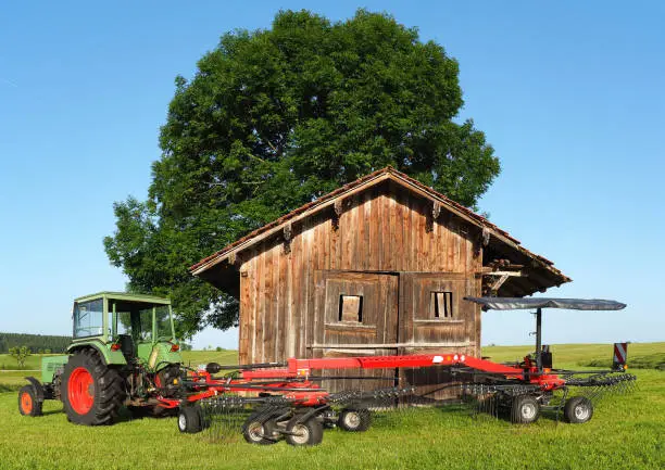 Marktoberdorf, Germany. Old Fendt tractor with trailed rotary tedder. Hay ready to be harvested. Wood storage in the countryside for tools. Hilly environment in Germany