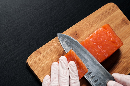 Hands cooks close-up. The chef cuts with a knife a red fish, smoked salmon on a wooden cutting board. Black wooden table on background.
