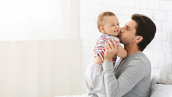 Happy paternity leave. Young man kissing his adorable baby son, enjoying babysitting, panorama with empty space