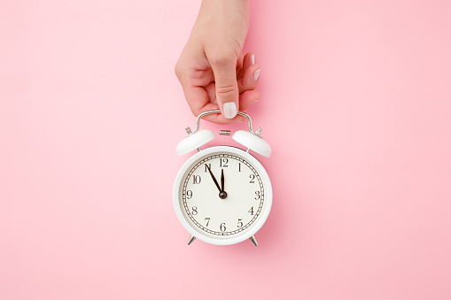 Woman hand holding white alarm clock on light pastel pink background. Time concept. Closeup. Top view.