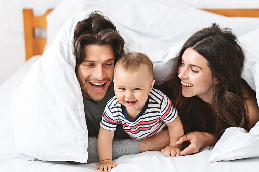 Family fun. Cheerful parents playing with baby son in bed, laughing together under blanket