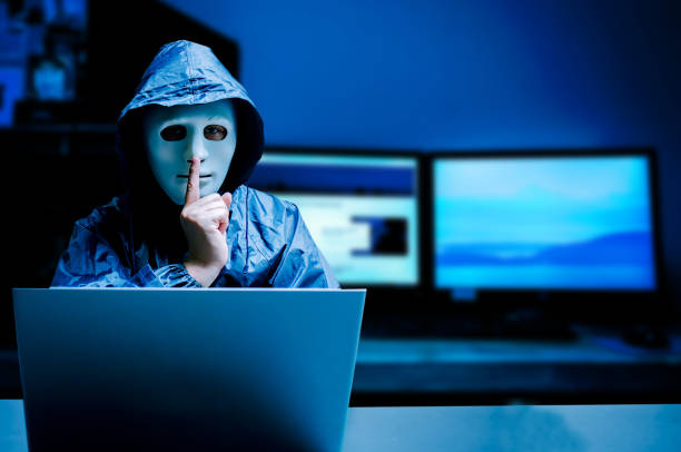 istock Anonymous computer hacker in white mask and hoodie. Obscured dark face making silence gesture on dark background, Data thief, internet attack, darknet and cyber security concept. 1165471764