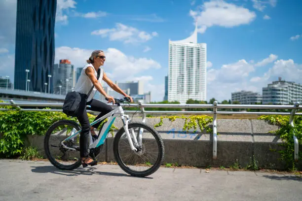 side view happy smiling 45 years old business woman cycling on electric bicycle outdoors on sunny summer day in european city, office buildings blurred in background