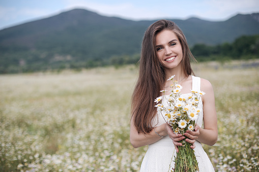 Beautiful girl outdoors with a bouquet of flowers in a field of white daisies,enjoying nature. Beautiful Model with long hair in white dress having fun on summer Field with blooming flowers,Sun Light. Young Happy Woman on spring meadow, countryside