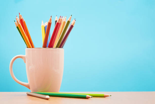 A white ceramic mug is full of rainbow colored pencil crayons; a few more lie next to it.
