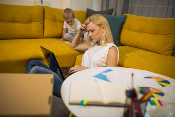 istock Young blonde mother working at home using laptop while her son is watching cartoons 1165485006