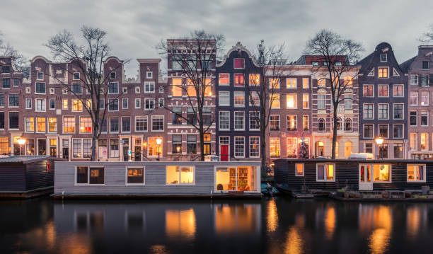 Traditional Dutch buildings and houseboats along the canals of Amsterdam, Netherlands Lights go on in homes for the night creating an artistic image. canal house photos stock pictures, royalty-free photos & images