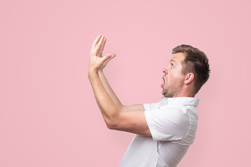 Oh my god. It is gorgeous. Shocked young man in white shirt with wide opened mouthlooking on his hands. Studio shot on pink background.