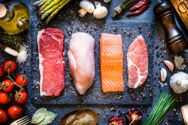 Top view of four different types of animal protein like a raw beef steak, a raw chicken breast, a raw salmon fillet and a raw pork steak on a stone tray. Stone tray is at the center of the image and is surrounded by condiments, spices and vegetables. Low key DSLR photo taken with Canon EOS 6D Mark II and Canon EF 24-105 mm f/4L