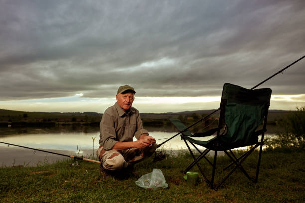 Old fisherman is fishing at the lake, has a fishing chair and fishing rods, the sky looks dark and cloudy