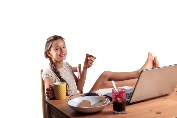 A happy smiling 10 years old school girl with an expressive emotional face sitting at the table with a laptop, eating bread with jam and holding a cup of tea isolated on white.