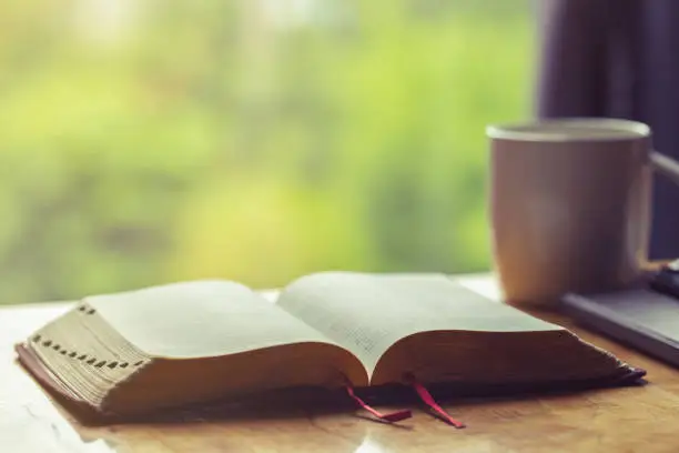 Photo of Open bible with a cup of coffee for morning devotion on wooden table with window light