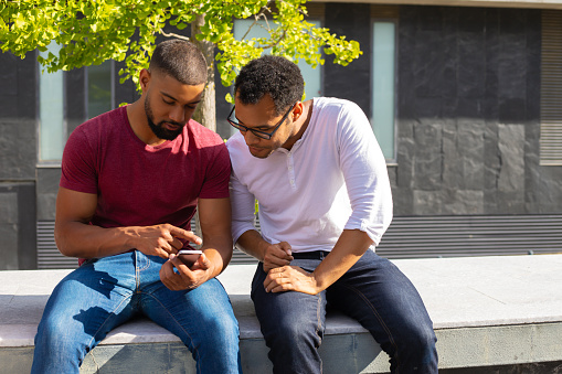 Serious male friends discussing online mobile app. Two men in casual sitting on parapet outside, staring at phone screen and talking. Communication concept