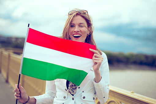 Happy young excited redhead woman holding Hungarian flag on bridge outdoors