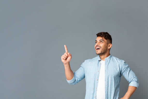 Excited Man Pointing Finger Upwards At Copyspace On Gray Background.