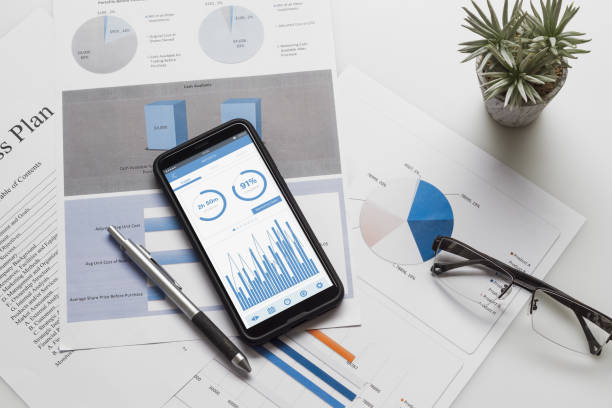 istock Top view of office desk table with graph chart on mock up smartphone on white desk table. Business management via mobile application concepts ideas. Mobk up mobile phone for your Advertisement. 1165525539