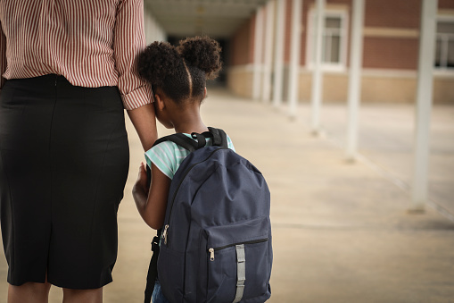 Elementary age, African American girl holds mom or teacher's hand before school begins.  She wears a backpack and clings to mom with uncertainty about starting school.
