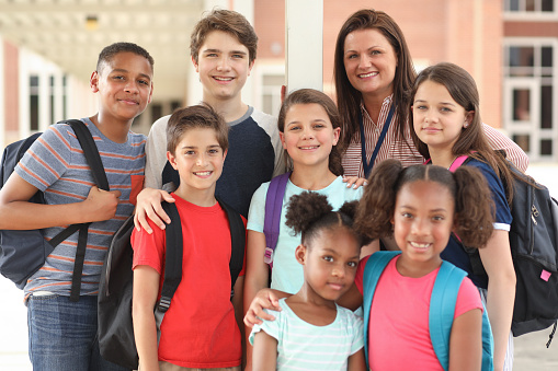Mixed age, multi-ethnic group of elementary and junior high age student friends hang out with teacher outside the school building before or after school. Caucasian, latin, and african descent.  Focus on Latin descent girl center.