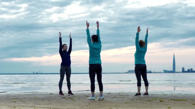 Mature yoga teacher explaining two adult female students how to do standing half forward bend with outstretched arms during practice near sea on beach in evening