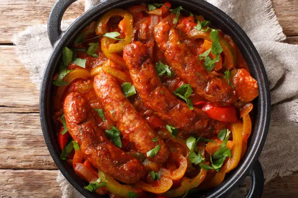 Photo of Traditional grilled sausages with multicolored peppers, onions and tomatoes close-up in a pan. horizontal top view