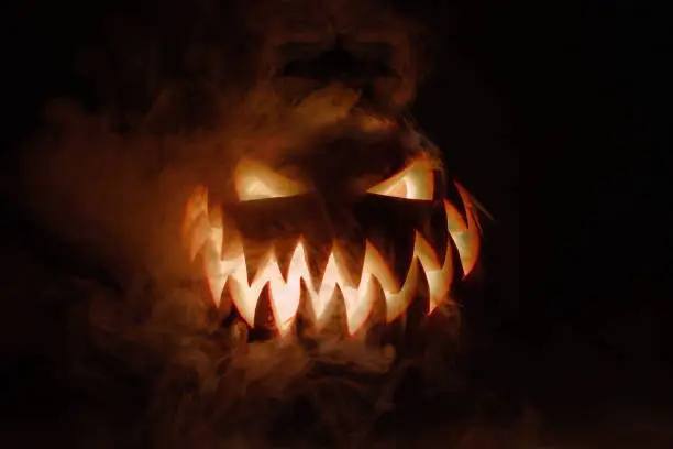 Photo of Fuming bright Jack-o'-lantern pumpkin on dark solid background. Glowing eyes and a terrible grin. Halloween minimal concept. Copy space. Desktop wallpapers
