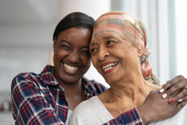 Courageous woman with cancer spends precious time with adult daughter A senior African American woman with cancer stands near a large wall of windows. She is embracing her adult daughter. They are laughing while resting their foreheads against each other. The women are full of hope for the future. adult age stock pictures, royalty-free photos & images