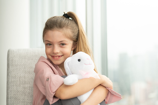 A cute elementary age girl is smiling into the camera while cuddling her stuffed animal. She is sitting in a chair by a large window.